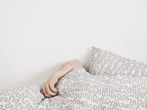 Against a  white background, a  light-skinned arm is visible, clutching a  white blanket with a gray doodle pattern on  it.  A  pillow  with the same pattern is visible just to the right of the arm.