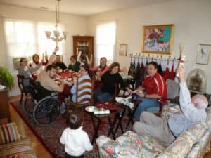 People gathered around a large table with others sitting on chairs and couch nearby.  A man at the table uses a wheelchair, and everybody is holding a Christmas Cracker up in the air.  A toddler stands beside the couch, his back to the camera.  Smiles are on all visible faces