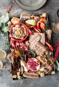 on a lightly colored wooden table, a large brown cutting board or tray is covered with festive food, including oranges, candy canes, pomegranates, gingerbread, and more.  