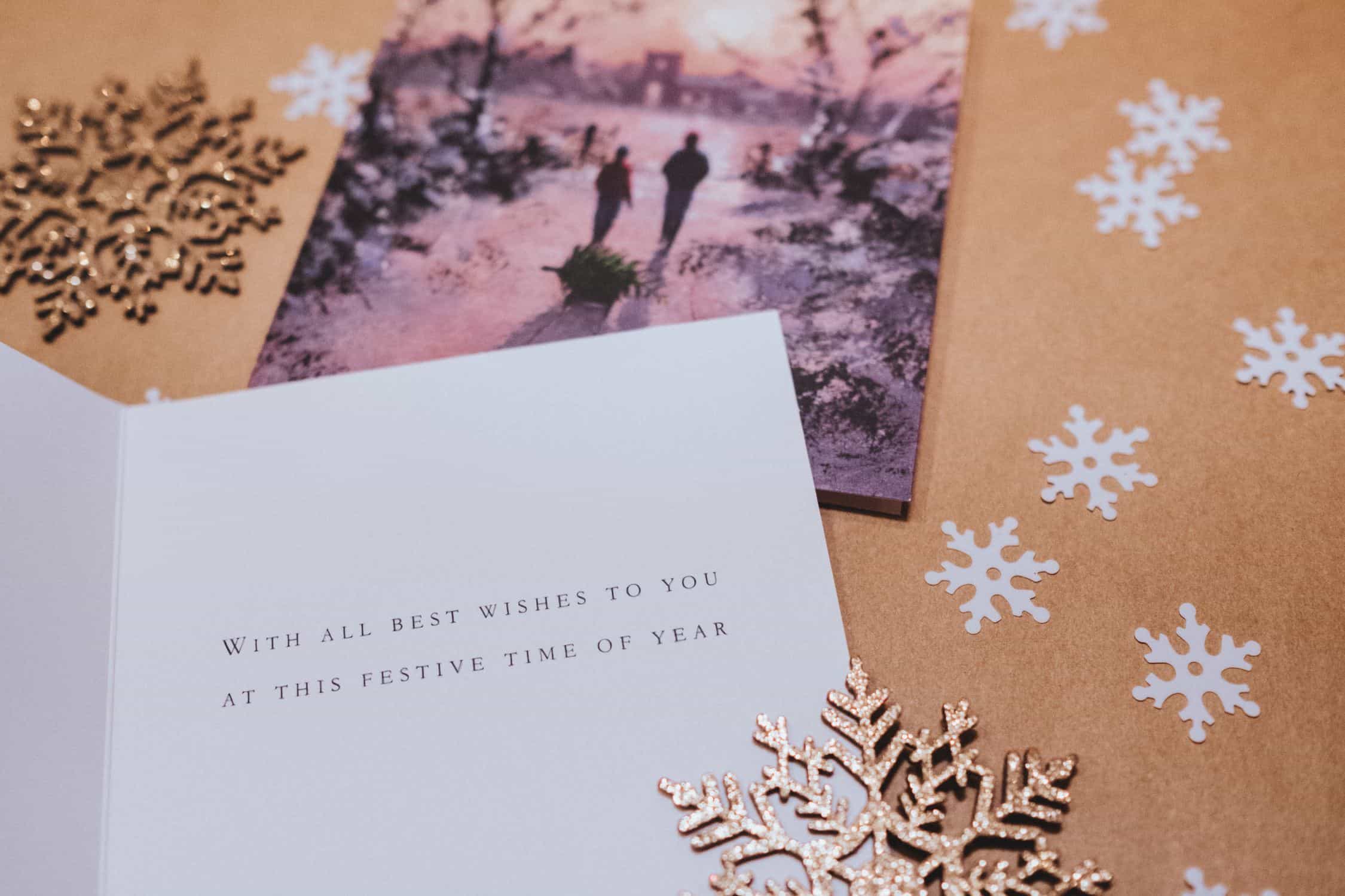 on a golden-brown background, with stylized snowflakes, two large holiday cards are layed out, as well as two golden snowflake decorations. One card is closed and picutes two people walking side by side in a winter wonderland. the card in front of it is open and shows two lines of text, though the words aren't quite readable