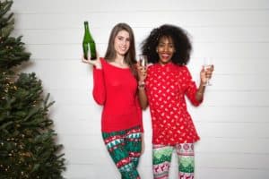 Two women, dressed in red tops and christmas-themed leggings raise glasses with the one on the left holding up a large green bottle.  The woman to the left is white and the woman to the right is black, both are smiling.  An undecorated pine tree is on the far left of the picture and they are standing against a white cinderblock wall.  
