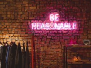 An indoor brick wall, with a coat hanger to the left and a set of shelves to the right.  The center of the image is a pink neon sign that says "Be Reasonable" in all caps.