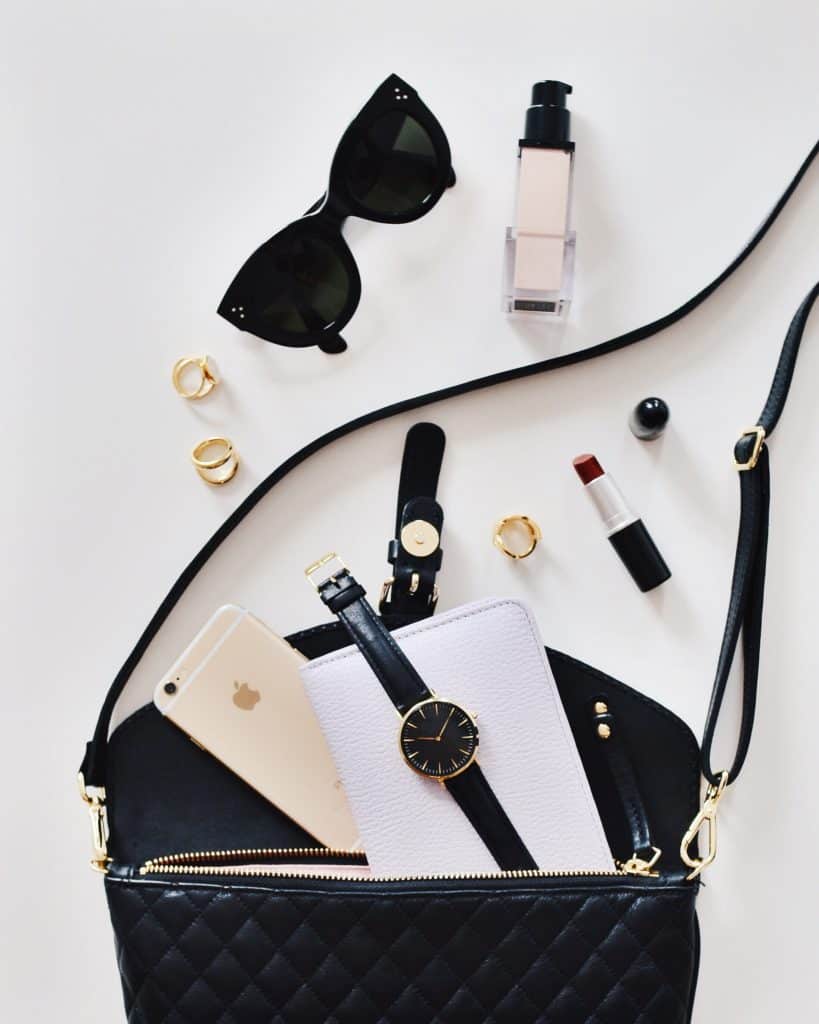 a black leather purse lies on a white surface.  Strewn around it are various objects often found in purses, like sunglasses, a watch, a cell phone, a planner, lipstick, earrings, and lotions.  