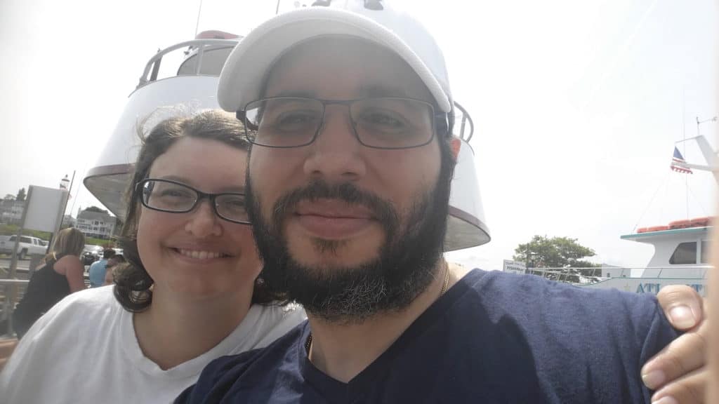 a Hispanic man with black hair, a beard and glasses and wearing a white hat.  Slightly behind him is a white woman with brown shoulder-length hair wearing glasses.   She is smiling, and he has a more neutral expression on his face.   They are on a fishing boat.