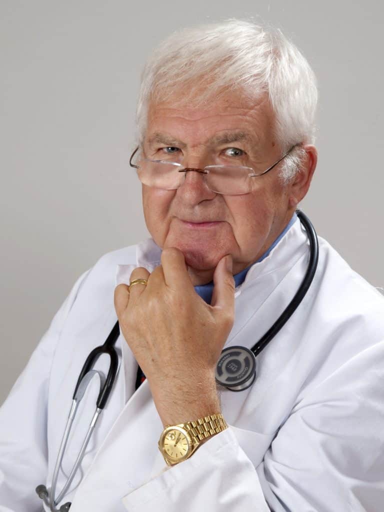 older white man dressed as doctor. He has white hair and glasses, and is looking over the top of the glasses into the camera