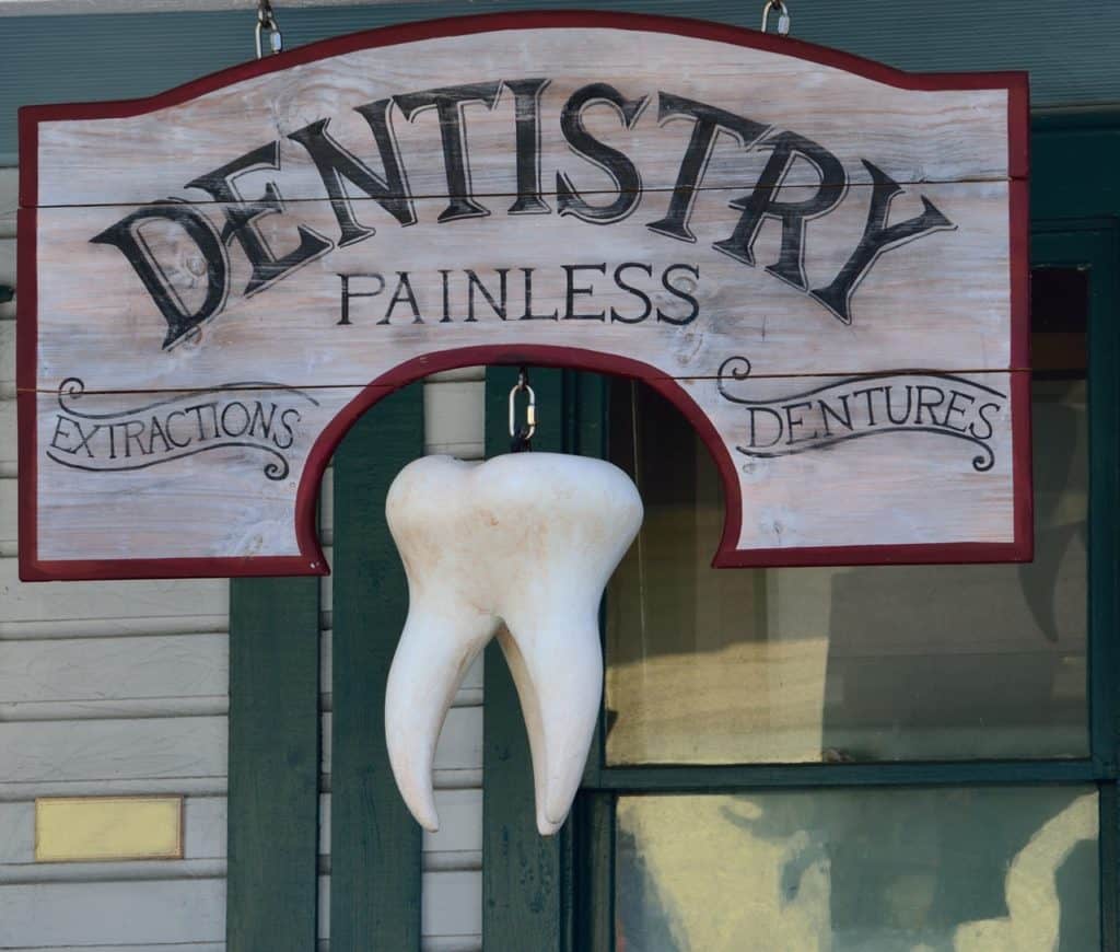 An old-fashioned sign hung in front of a building.  The sign has a 3-dimensional representation of a tooth(including 3 roots) hanging off of it and reads "Dentistry", with "Painless" below it in smaller letters.  the left side of the sign reads "extractions" and the right "dentures". 