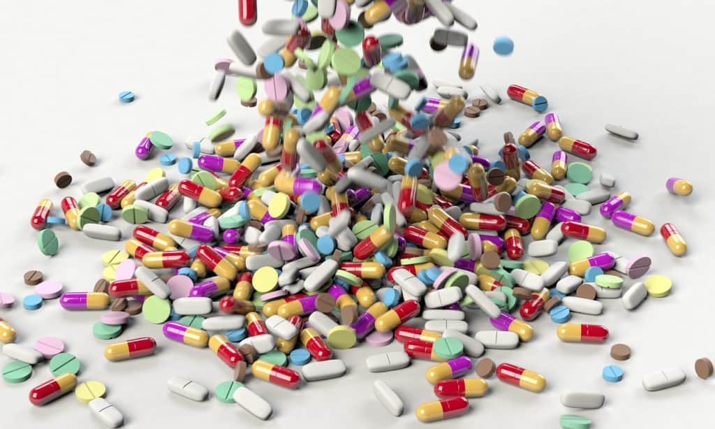 On a white background, a huge variety of medications in various shapes, sizes, and colors.  It is an action shot, with medications still being dropped onto the white surface. 