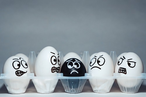 Eggs in an egg carton. All are white except for a single black egg in the center. All eggs have faces drawn on them - with the black egg looking frightened, and the surrounding white eggs looking varying degrees of angry or disapproving and appearing focused on the black egg