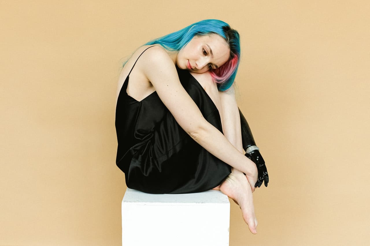 woman in black sleeveless dress sitting on a white surface. She has blue hair with a pink streak on the right side, and her left arm is prosthetic.