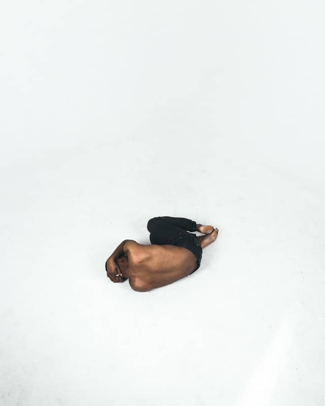On a large white space, a black man in black  pants curls up in close to the fetal position.  His head is bowed down and covered by his  right arm, with his left arm tucked under his body.  He appears to be making himself as compact as possible.