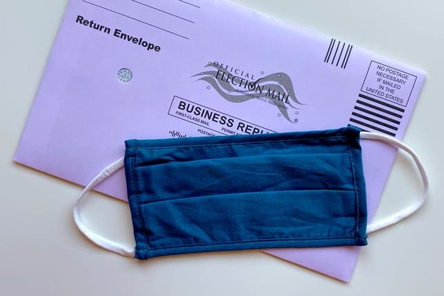 an official US ballot envelope, purple, with a blue cloth facemask above it.