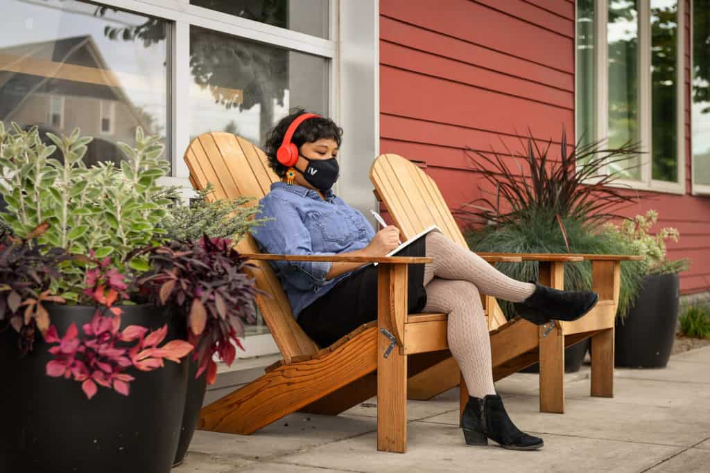 A Filipinx woman with a filtering face mask sits outside a cafe and writes in a notebook. She is wearing headphones and in front of a window, with potted plants all around.