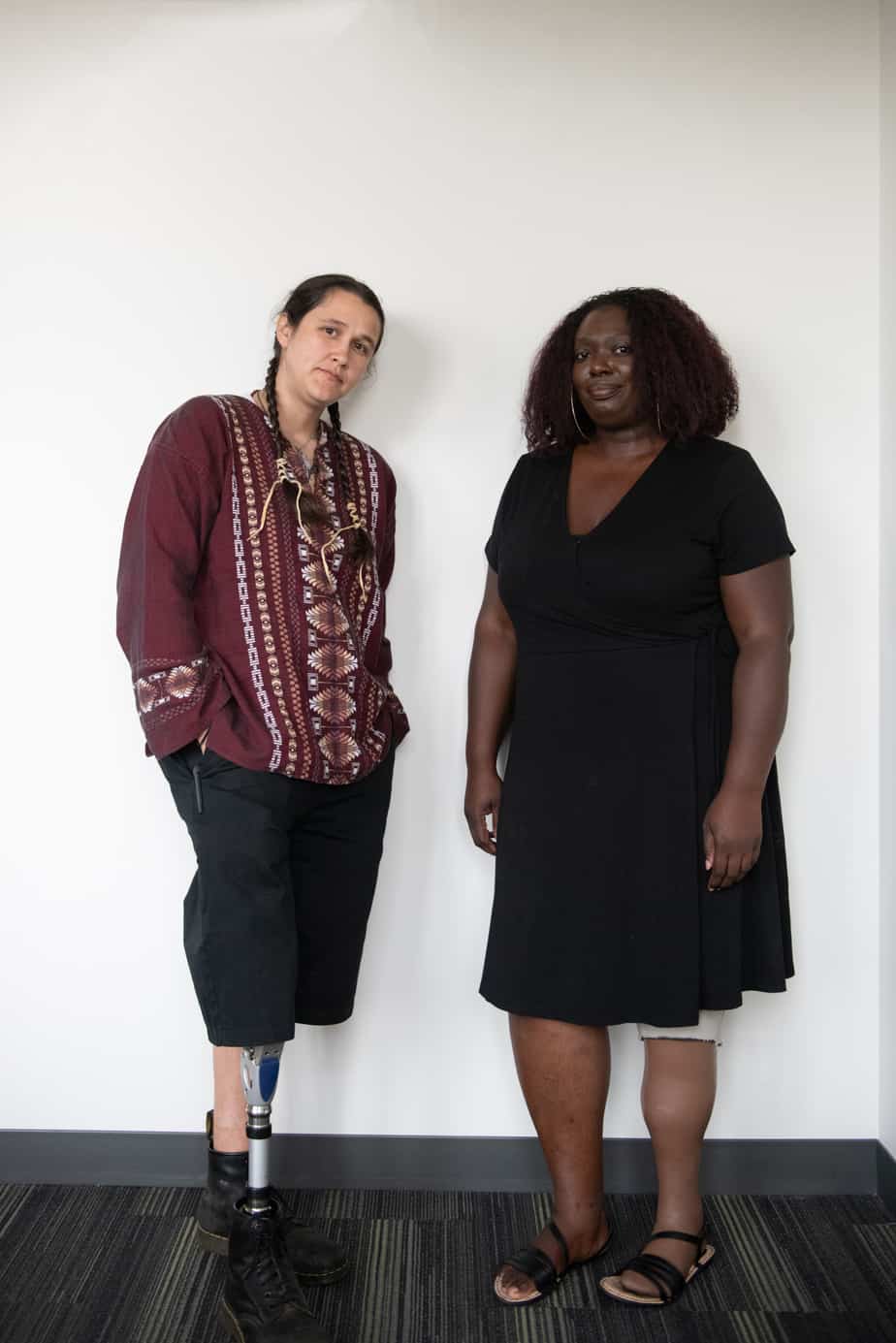 An Indigenous Two-Spirit person and a Black woman stand in front of a white wall with neutral expressions. They are both wearing prosthetic legs.