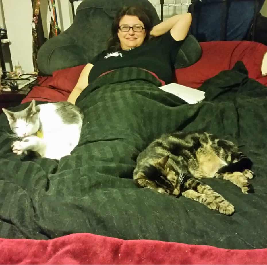 Alison is lying in bed, with a black comforter over her. Rorschach is cleaning himself while leaning against her right side, just below her hip, while Nigel is sprawled out a bit just to the left of her legs