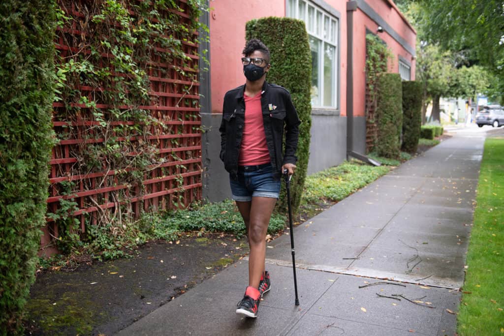 A Black non-binary person with a filtering face mask walks down a neighborhood street with one hand in their pocket and the other hand on their cane. They have a short mohawk and are wearing a jacket, shorts, tennis shoes, and glasses.