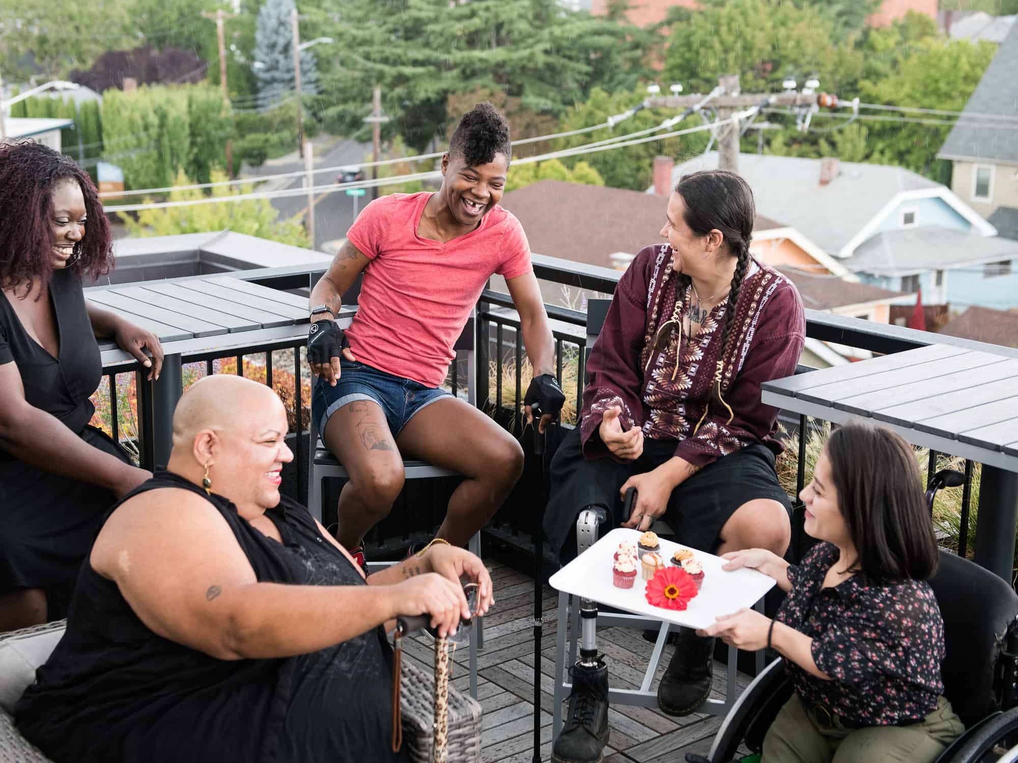 A South Asian person in a wheelchair brings mini cupcakes out on a platter to four other excited disabled people of color. They are all sitting around a rooftop deck.