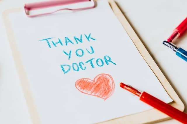 On a clipboard, there's a piece of white paper with crayon writing on it.  The blue writing reads 'Thank you doctor' and a large, red heart is drawn underneath and colored in.