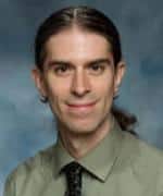 picture of a white man with long brown hair pulled back in a ponytail. The view is cropped at the shoulders, and he's wearing a gray collared shirt and a dark tie. He has a hint of a smile on his face in this formal picture