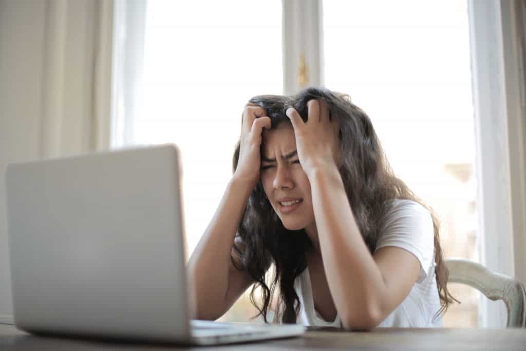 woman with long brown hair sits at a table with a laptop computer in front of her.  She is hunched in front of the keyboard and has her head in her hands and looking upset and frustrated.