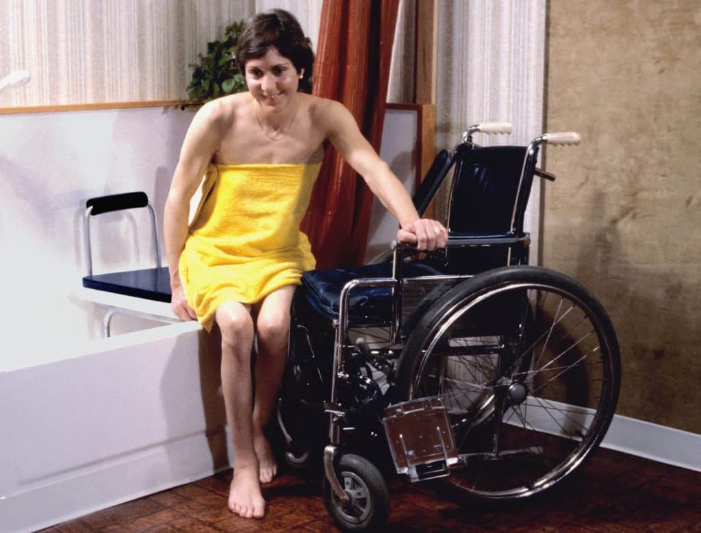 female wheelchair user wrapped in a yellow towel sits on a shower bench next to her wheelchair
