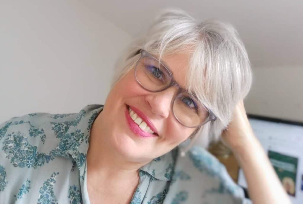 A white woman with short gray hair and wearing dark-rimmed glasses tilts her head to the right, leaning it against her right hand and smiles into the camera. She's in a close-cropped photo that shows her to chest height and she's wearing a grey-blue collared shirt with paisley-shaped collections of turqoise blue tiny flowers dotting it. The photo feels friendly and inviting, like she's just asked you a question and is attentively awaiting your answer.