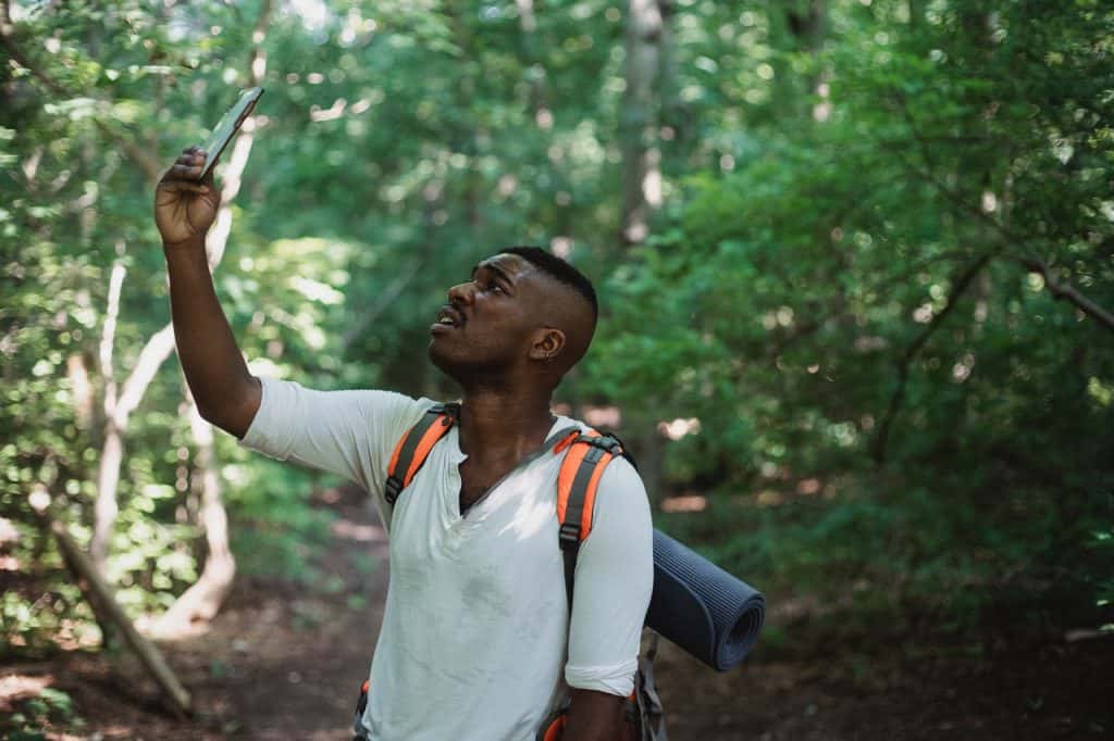 black man holds up cell phone, apparently searching for a signal, while standing in a forest