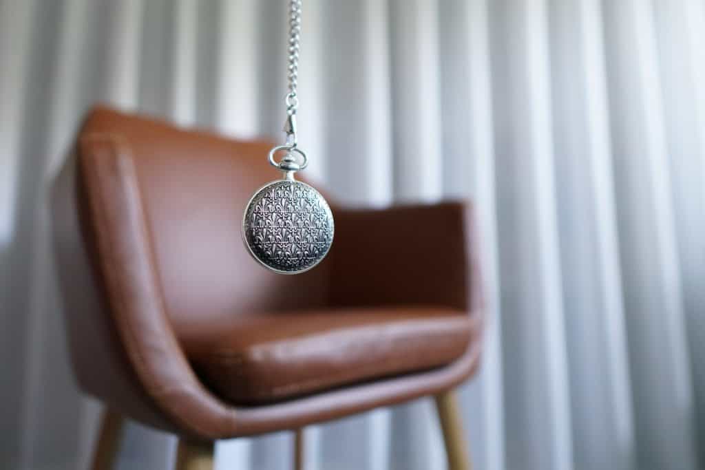 A leather chair sits against a white curtain.  In the foreground, a silver round pendulum dangles