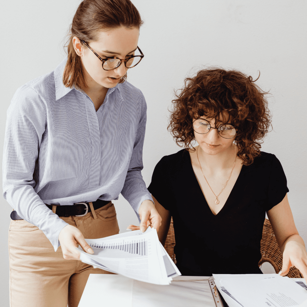One white woman with curly brown hair and glasses sits at a table, looking at papers, while another white woman, with long straight brown hair pulled back in a ponytail stands by her, showing her additional documents. Both are focused on the work in front of them.