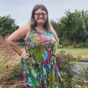 Alison(white woman with shoulder-length brown hair with white highlights and dark glasses) stands surrounded by greenery. She is smiling and has her hands on her hips, wearing a multicolored(primarily blues, greens, and purples) patchwork dress.