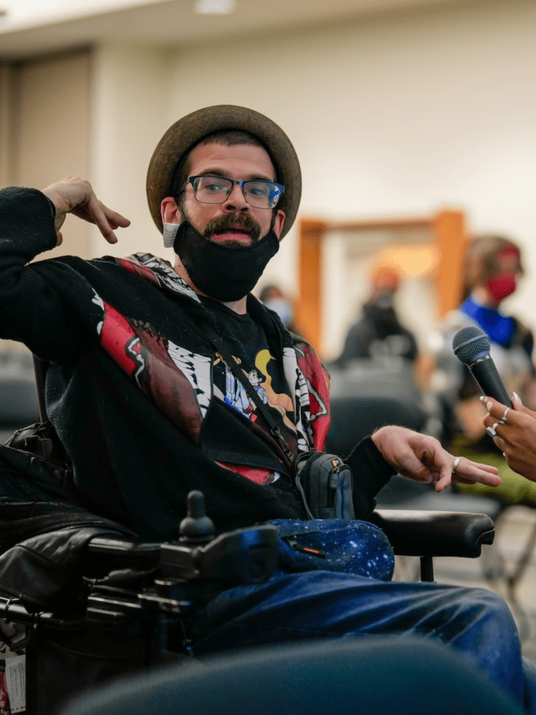 Light-skinned man in wheelchair with an anxious or concerned look on his face raises his hand. He is a wheelchair user with glasses, dark hair, and a mustache, with a black mask covering his chin. He is wearing a black shirt with a multicolored pattern on it and blue jeans.
