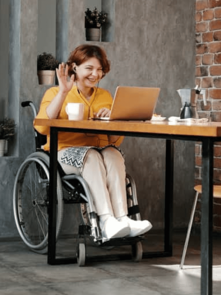 woman in a wheelchair has her computer open and is waving to somebody on the screen