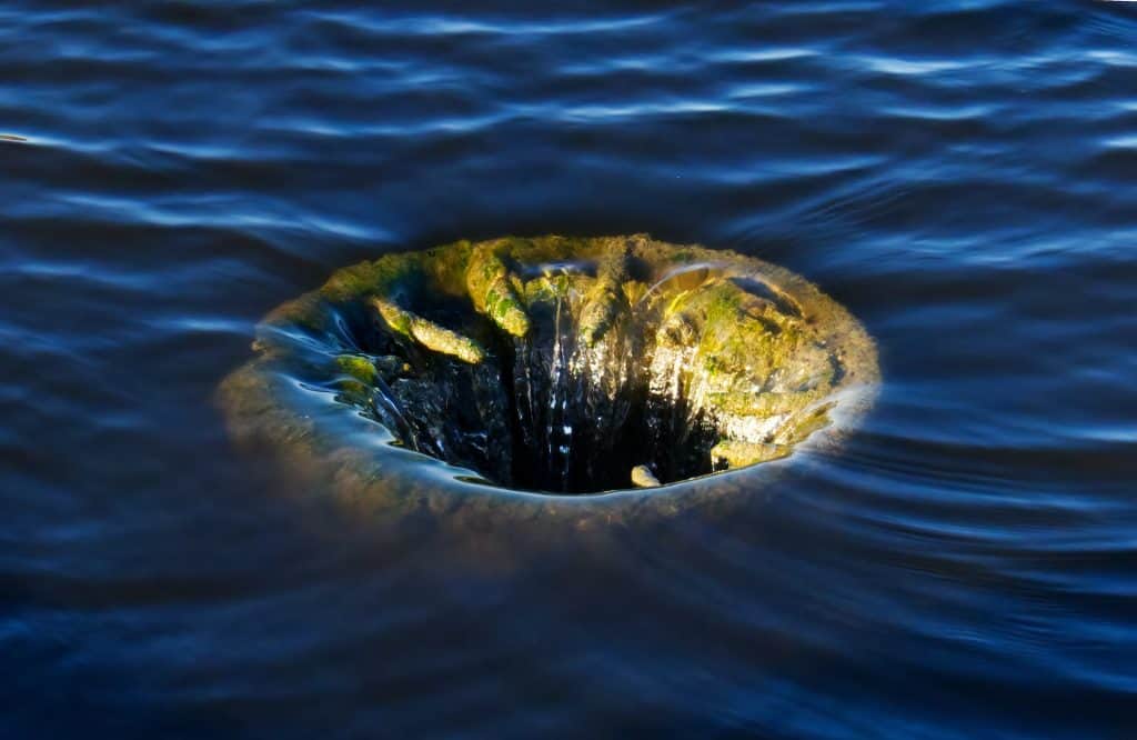 In a closeup of a body of water, the central focus is a rusted hole that the water is pouring  into. 