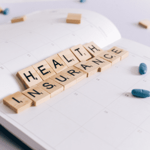 A monthly planner lies open on a white table. the words "health insurance" are spelled out across the fold in the planner using wooden scrabble tiles. more tiles lie scattered across the planner and table, face down. Blue pills are also visible scattered across the planner and table.