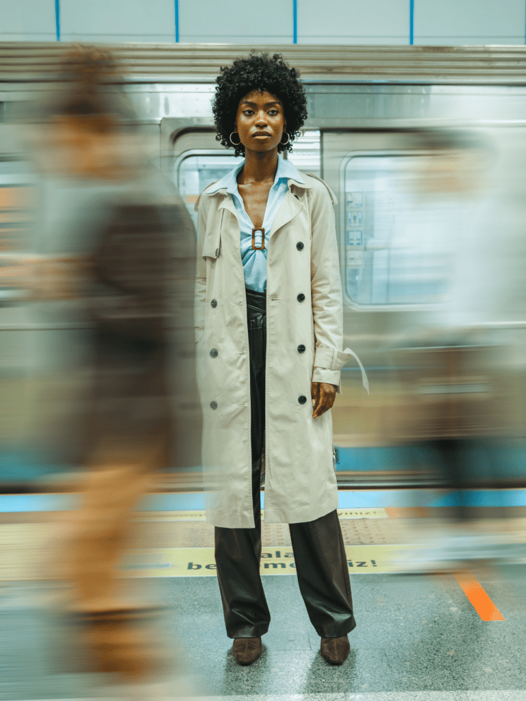 Black woman in trenchcoat stands, looking slightly lost, with a blurred train behind her and people walking past her, all blurred to imply motion.